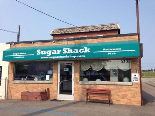 Exterior of Sugar Shack bakery featuring a green awning, sign with "Cupcakes, Cookies, Brownies, Pies," and a welcoming entrance.