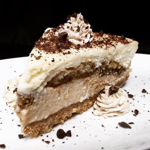 A slice of layered dessert with creamy filling, topped with whipped cream and chocolate shavings on a white plate.