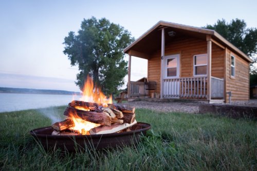 A cozy cabin by the lake with a campfire in the foreground, surrounded by grass and trees at dusk.