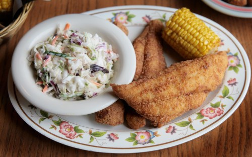 A plate featuring fried fish fillets, coleslaw, and a corn on the cob, garnished with a floral border.