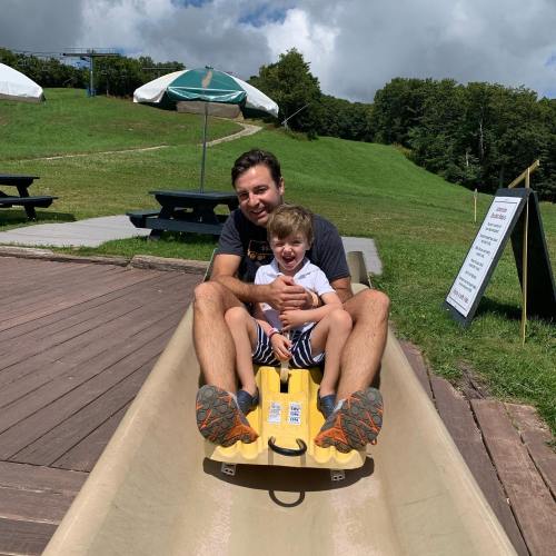 A man and a young boy sit together on a slide, smiling, with green hills and picnic tables in the background.