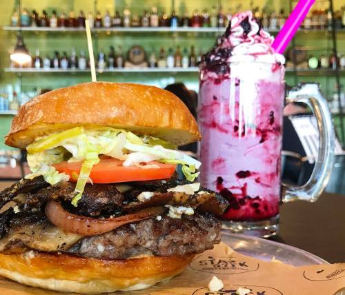 A juicy burger with lettuce, tomato, and grilled onions, alongside a colorful milkshake topped with whipped cream.