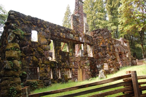 Ruins of a stone building surrounded by trees, with moss-covered walls and a wooden fence in the foreground.