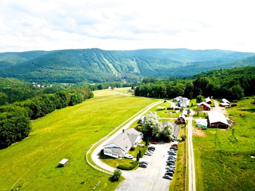 Aerial view of a green valley with a road, buildings, and mountains in the background under a bright sky.