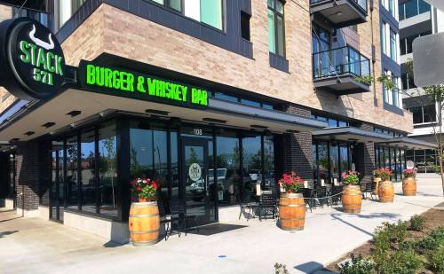 Exterior of Stack 571 Burger & Whiskey Bar, featuring large windows, outdoor seating, and decorative flower barrels.