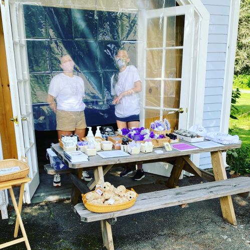 Two people wearing masks stand behind a table with various products, set up outside a building with open doors.