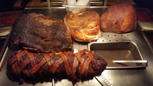 A variety of smoked meats, including brisket, a seasoned loaf, and a bacon-wrapped dish, displayed on a grill tray.