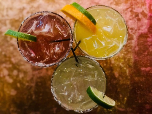 Three colorful cocktails with ice, garnished with lime and orange slices, on a textured background.