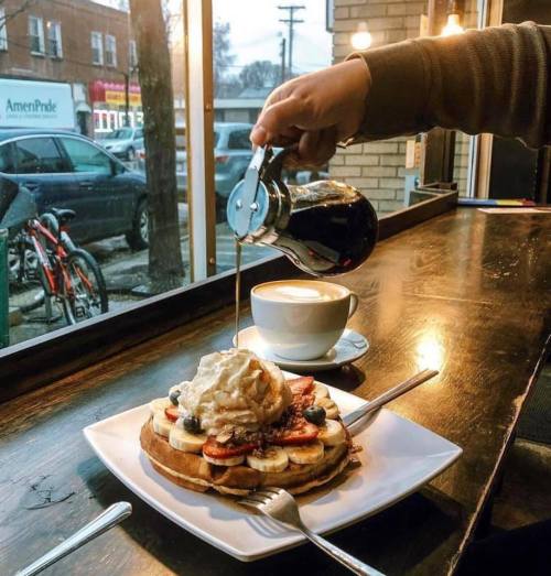 A hand pours syrup over a stack of pancakes topped with bananas, blueberries, and whipped cream, beside a cup of coffee.