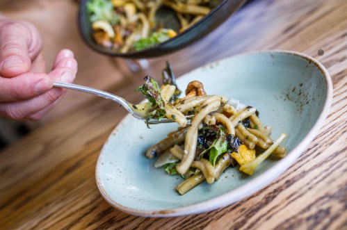 A hand holding a fork with a serving of noodles and vegetables on a light blue plate, set on a wooden table.