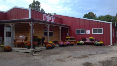 A red barn-style building with a sign reading "Apple Hut," surrounded by colorful flower pots and a gravel path.