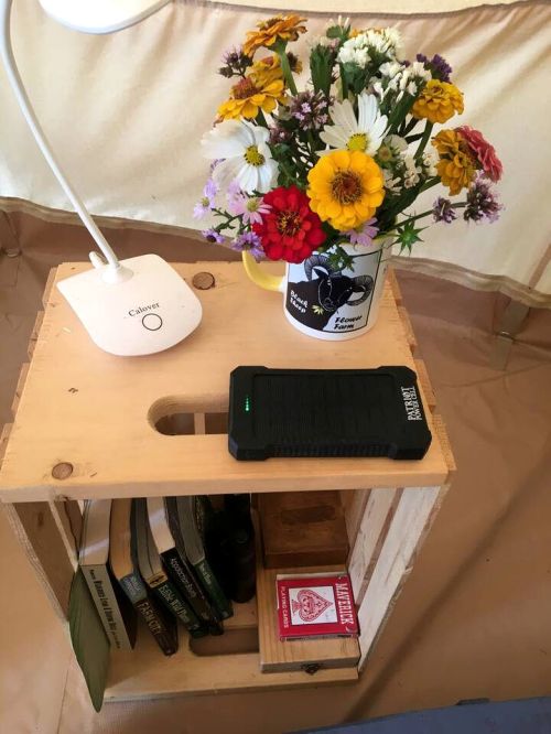 A wooden crate table holds books, a portable charger, and a colorful flower arrangement in a mug, with a lamp nearby.
