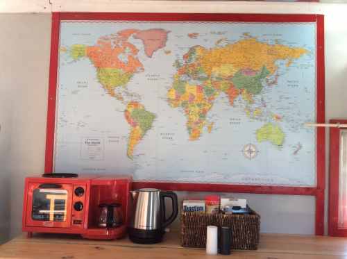A world map hangs on a wall above a wooden table with a red coffee maker, kettle, and a woven basket.
