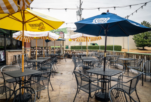 Outdoor dining area with black tables and chairs, colorful umbrellas, and a cloudy sky in the background.
