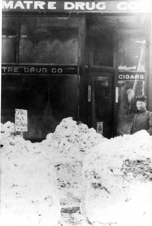 A historic black-and-white photo of a snowy street with a drugstore and a man standing beside large snowdrifts.