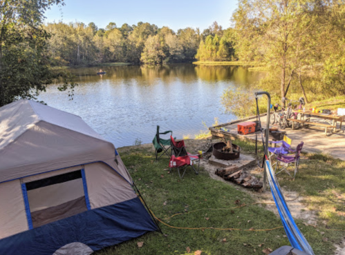 A campsite by a calm lake, featuring a tent, chairs, a fire pit, and a picnic table surrounded by trees.