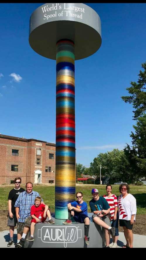 A group of seven people poses beside the World's Largest Spool of Thread, featuring colorful threads and a clear blue sky.