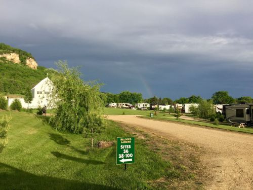 A campground with RVs, green trees, and a sign for sites 36-100 under a cloudy sky with a hint of a rainbow.
