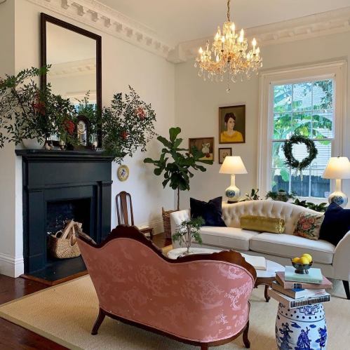 Cozy living room with a pink sofa, chandelier, plants, and a fireplace, featuring elegant decor and natural light.