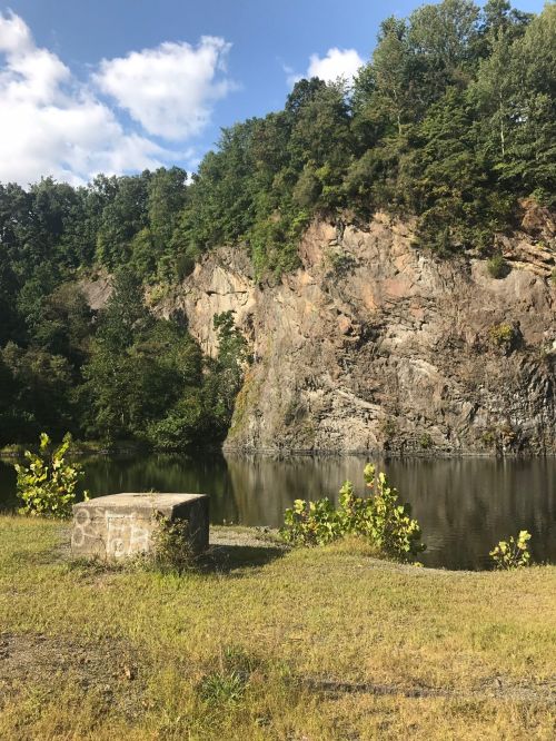 A rocky cliff rises above a calm lake, surrounded by trees under a blue sky with scattered clouds.