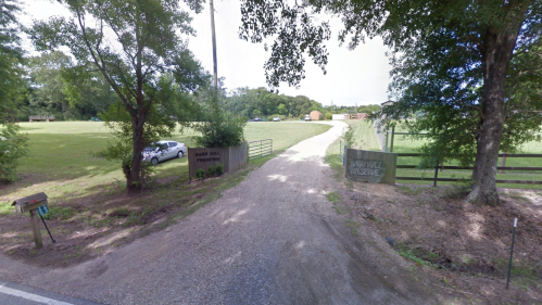 Gravel driveway leading to a park entrance with signs, surrounded by trees and open fields.