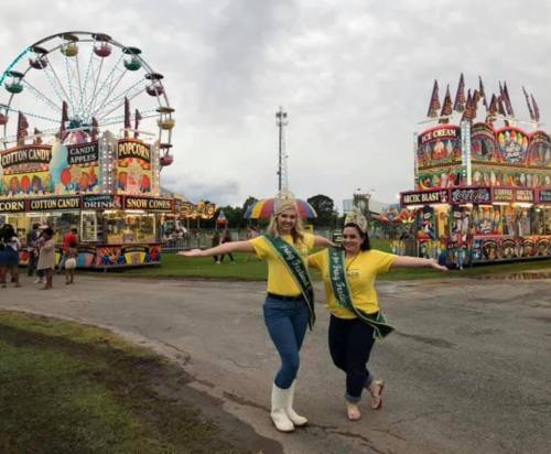 Two women in yellow shirts and sashes pose joyfully at a fair, with colorful rides and food stalls in the background.