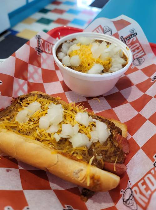 A hot dog topped with chili, onions, and cheese, alongside a bowl of chili with onions and cheese, served in a red basket.