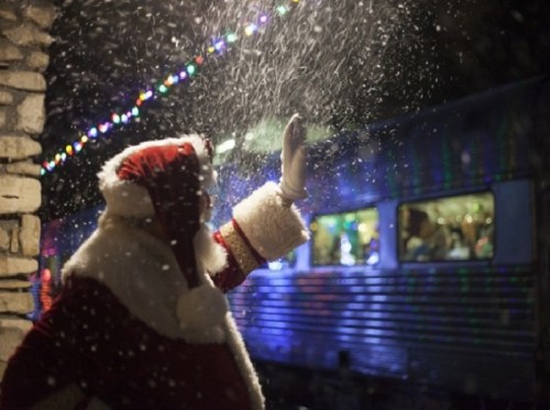 Santa Claus waves in the snow, with colorful lights and a train in the background.