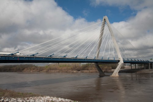A modern cable-stayed bridge spans a river under a cloudy sky, with greenery along the riverbank.