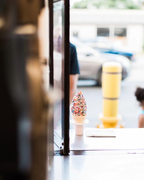 A colorful ice cream cone with sprinkles sits on a counter, viewed through an open window.