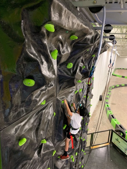 A child climbs a rock wall with green holds, wearing a safety harness and helmet, in an indoor climbing facility.