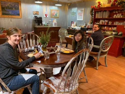 Three people sit at a rustic table in a cozy café, enjoying breakfast in a warmly decorated space.