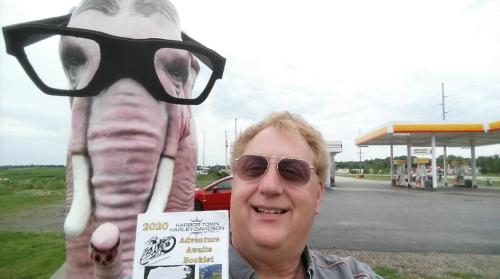 A man takes a selfie with a large elephant statue wearing glasses at a gas station.
