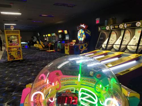 A vibrant arcade filled with various games, including skee-ball and a colorful prize wheel, under dim lighting.