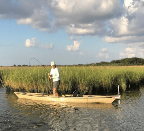 A person fishing from a small boat in a marshy area with tall grass and a cloudy sky.