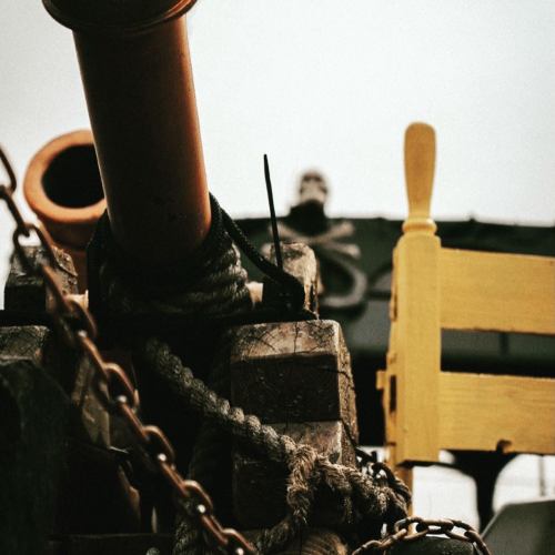 A close-up of a ship's cannon, ropes, and a yellow chair against a cloudy sky.
