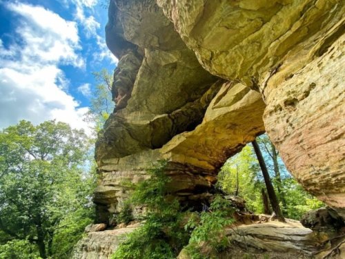 A large rock formation with a natural arch, surrounded by lush greenery and a bright blue sky with clouds.