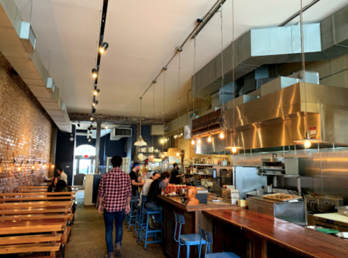 A modern restaurant interior with wooden tables, a bar area, and an open kitchen, featuring industrial decor and warm lighting.