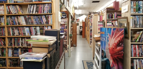 A narrow aisle between tall shelves filled with books and DVDs in a cozy bookstore.