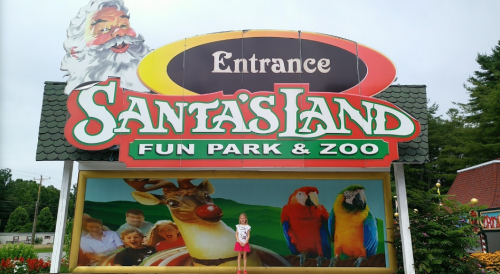 A child stands in front of the colorful entrance sign for Santa's Land Fun Park & Zoo, featuring Santa and animals.