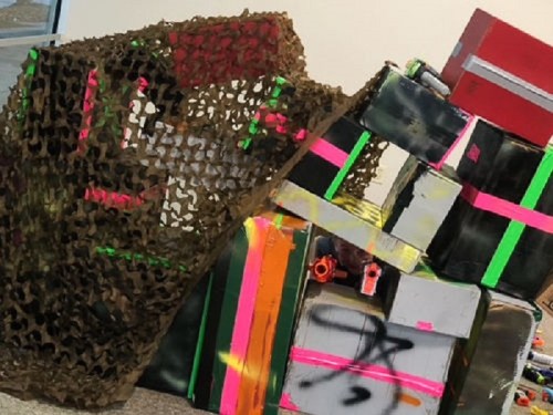 A pile of colorful boxes covered with a camouflage netting, featuring bright neon markings and graffiti.