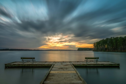 A serene lake view at sunset, featuring a wooden dock with two benches and dramatic clouds in the sky.
