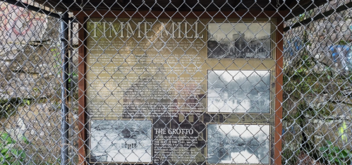 Sign about Timmer Mill, featuring historical photos and information, enclosed in a chain-link fence.