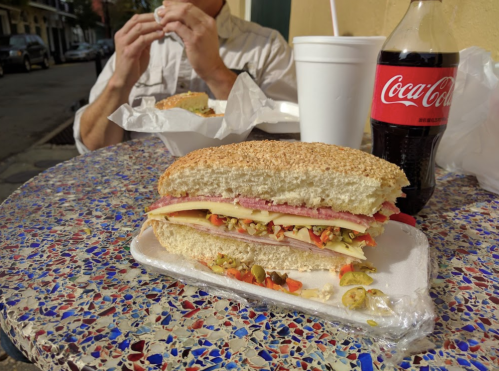A close-up of a sandwich with layers of meat and cheese, next to a Coca-Cola on a colorful table.
