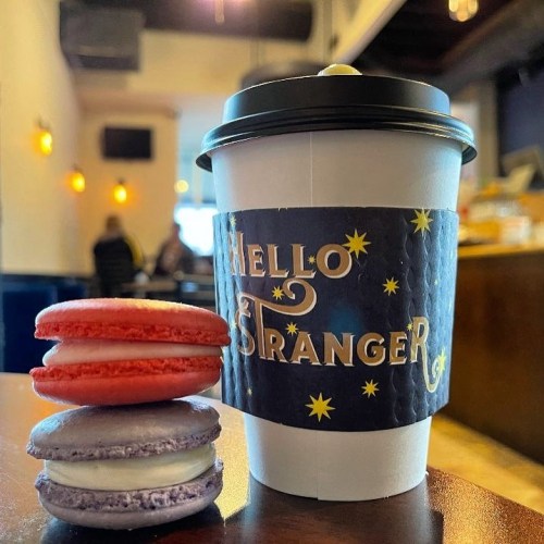 A coffee cup with a "Hello Stranger" sleeve next to red and purple macarons on a wooden table.