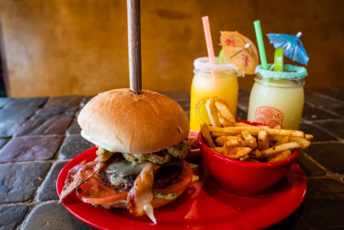 A burger with bacon, lettuce, and tomato, served with fries and colorful drinks with umbrellas on a red plate.