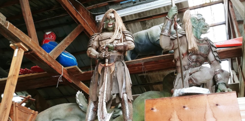 Two large, detailed statues of warriors with green skin and armor, displayed on a wooden shelf in a rustic setting.