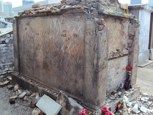 A weathered tomb with faded markings and scattered debris, surrounded by other graves in a cemetery.