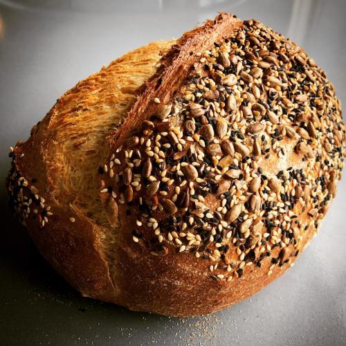 A round loaf of bread topped with a variety of seeds, including sesame and flax, on a gray surface.