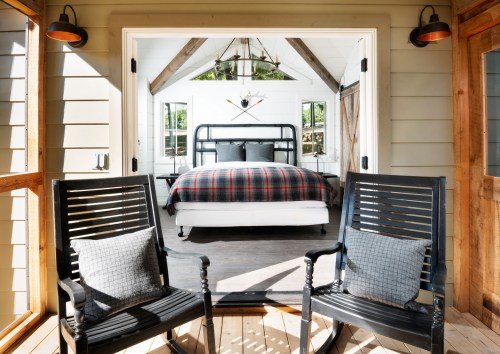 A cozy bedroom with a plaid bedspread, viewed through an open porch with two rocking chairs.